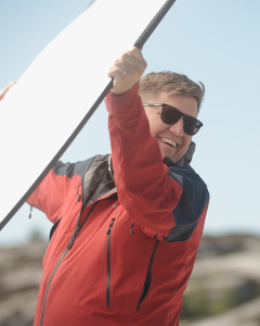 En person med solbriller og rød jakke holder opp en stor hvit solskjerm.