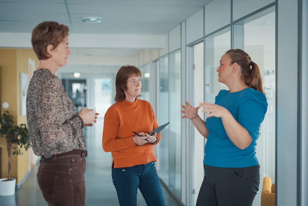 Tre personer står i en gang og diskuterer om de skal bytte regnskapsprogram. En person holder et nettbrett.