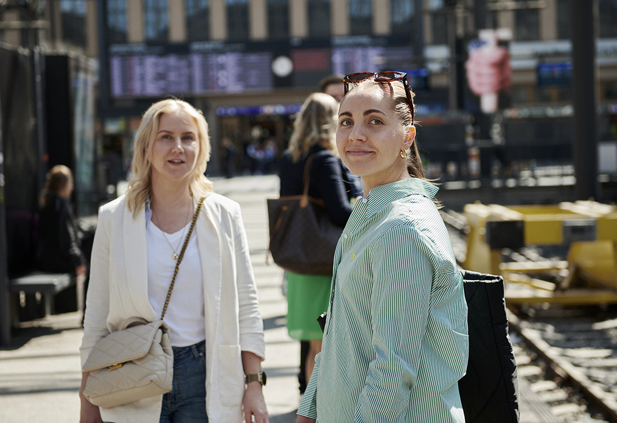 Reiseregning: to kvinner på jobbreise, er på togstasjonen, ser i kamera.