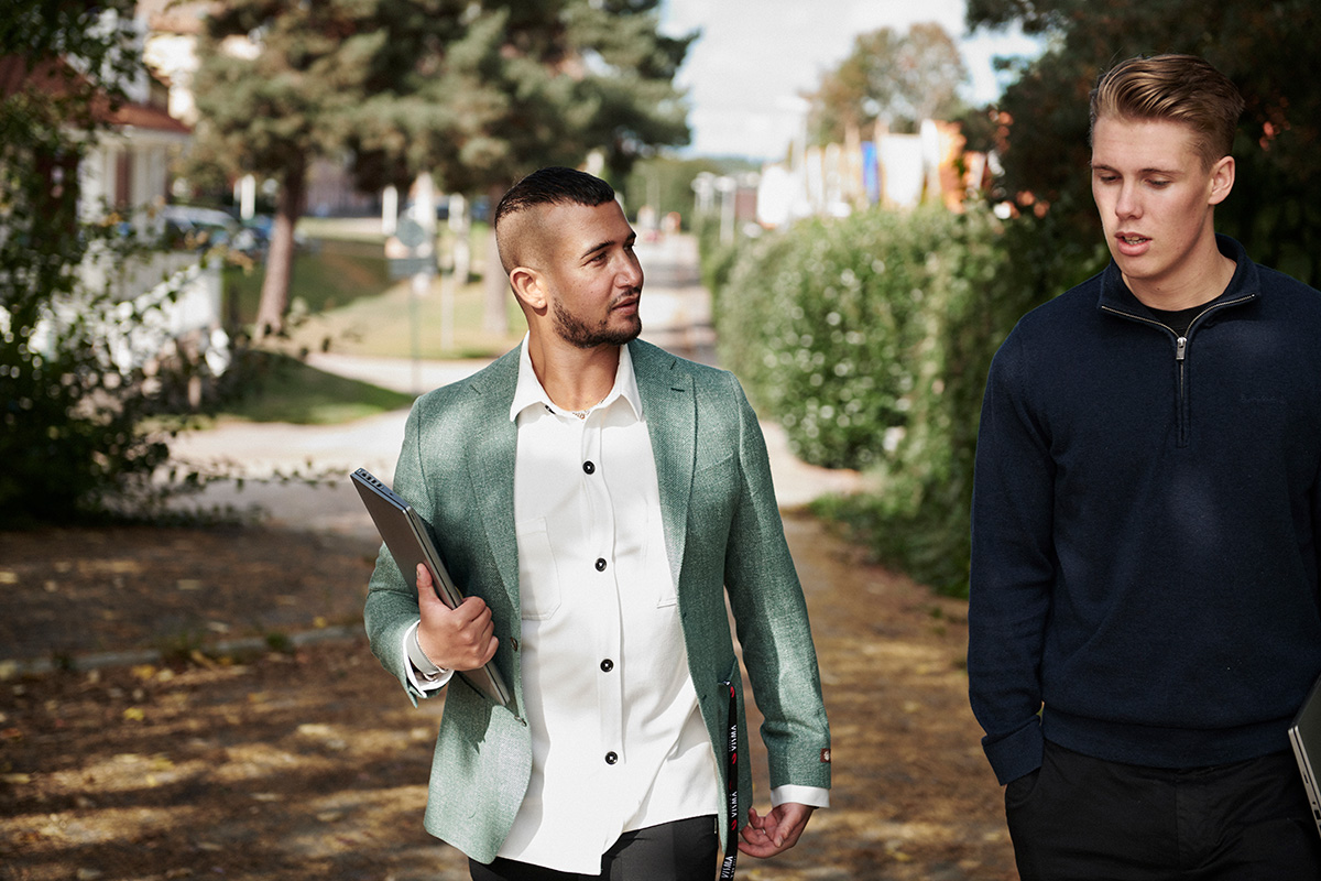 To menn som gikk ute på en gangsti, den ene i hvit skjorte og grønn blazer med en mappe merket «Forskuddsskatt» og den andre i en blå genser. Trær og bygninger er synlige i bakgrunnen.