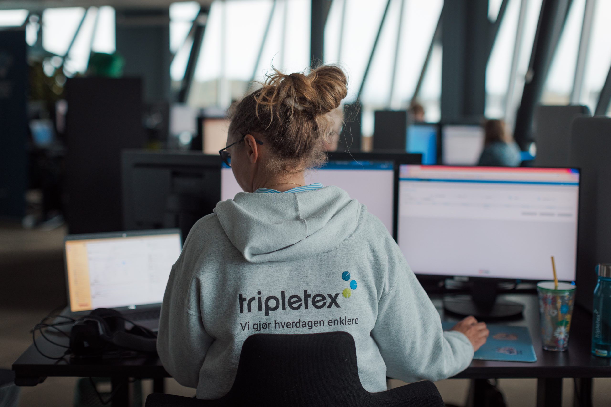 En person med "Tripletex"-hettegenser jobber ved et skrivebord med flere dataskjermer i et kontormiljø.