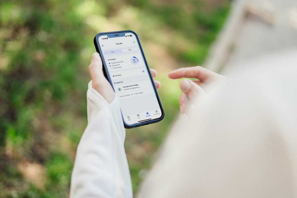 Feriepenger: En person holder en telefon utendørs med Tripletex-appen og registrerer ferie.