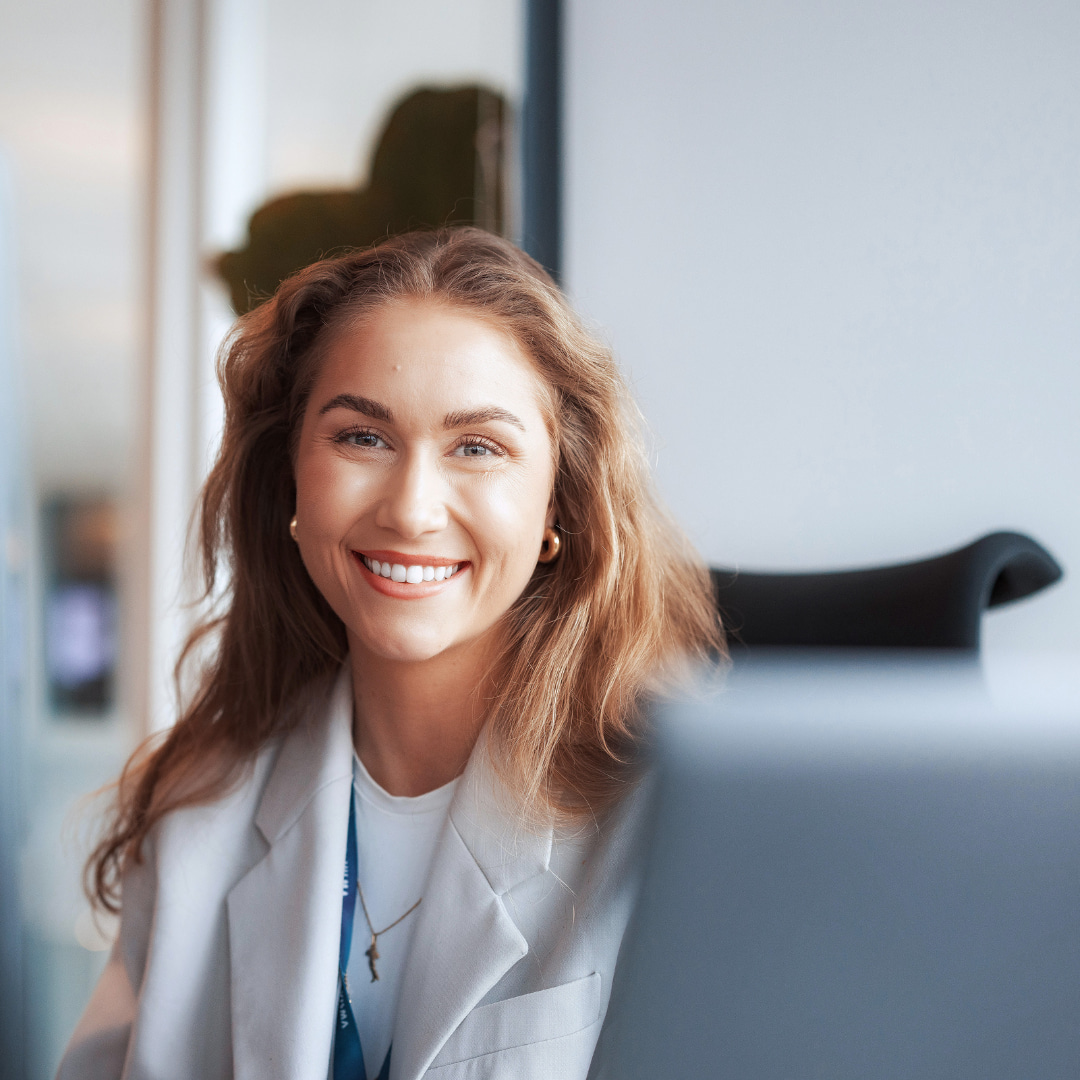 En kvinne med langt, bølget hår, iført en hvit blazer og topp med blå jeans, står innendørs og smiler til kameraet, klar til å presentere sitt nyeste prosjektstyringsverktøy.
