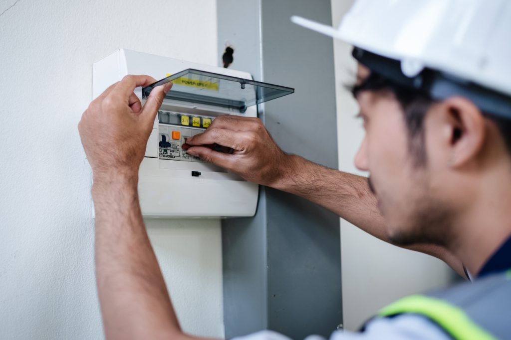 En person som har på seg en hjelm justerer brytere på et elektrisk panel montert på en vegg. Kontrollskjema brukes.