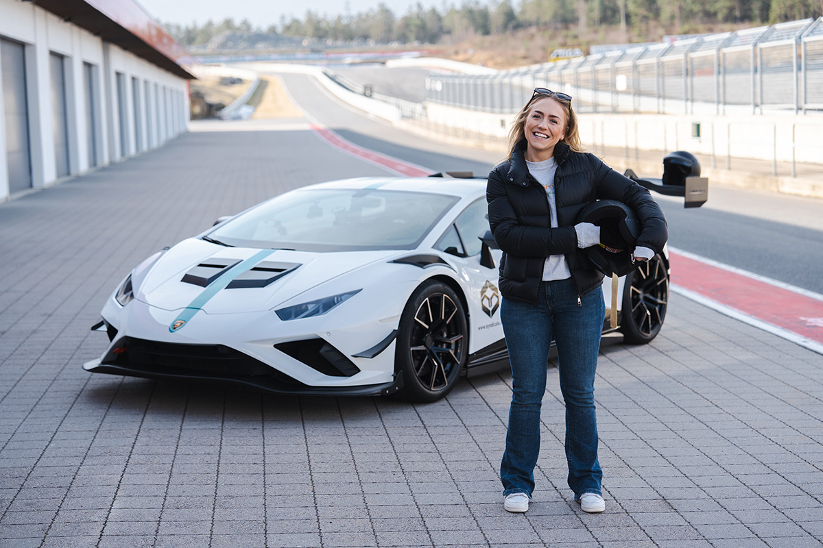 En kvinne i svart jakke og jeans står og smiler foran en hvit sportsbil på en racerbane. Er Tripletex raskere enn en Lamborghini?
