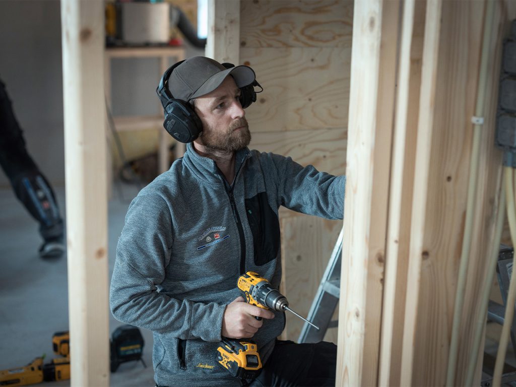 En håndverker med øreklokker bruker en boremaskin mens han jobber på et reisverk.
