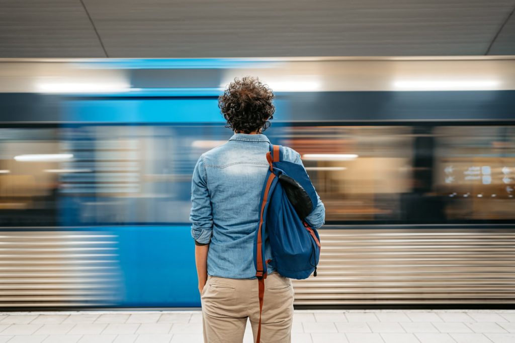 En person med en blå ryggsekk venter på en t-baneplattform mens et blått tog passerer.