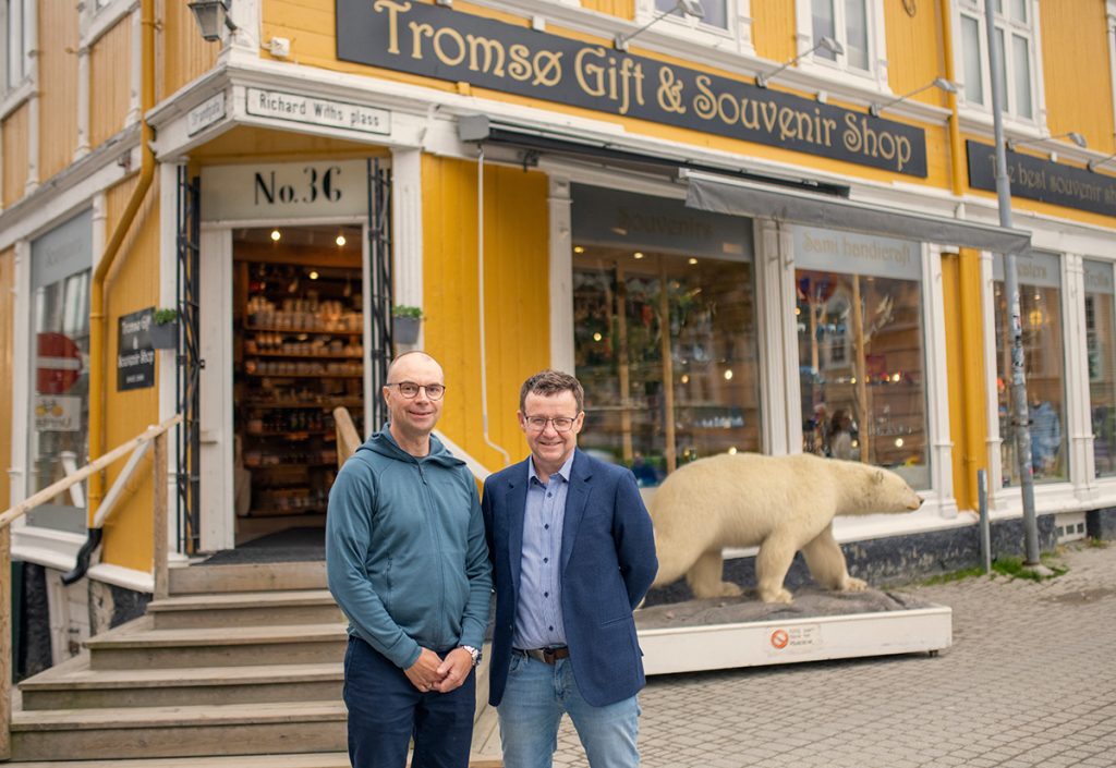 To menn som står utenfor Tromsø Gift- og Souvenir Shop med en utstoppet isbjørn synlig gjennom butikkvinduet. Snakker om Trygg-Økonomi.