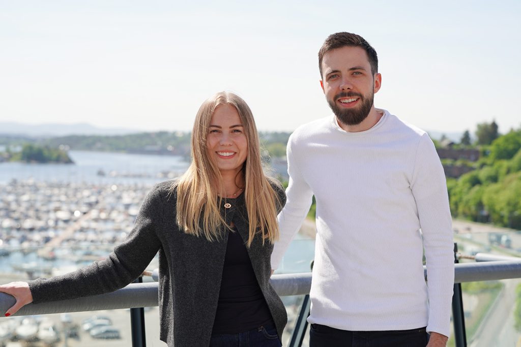 To personer står utendørs på en balkong med utsikt over en marina og snakker om utviklerglede. Kvinnen til venstre har på seg en grå genser, og mannen til høyre har på seg en hvit genser. Begge smiler med grønt i bakgrunnen.