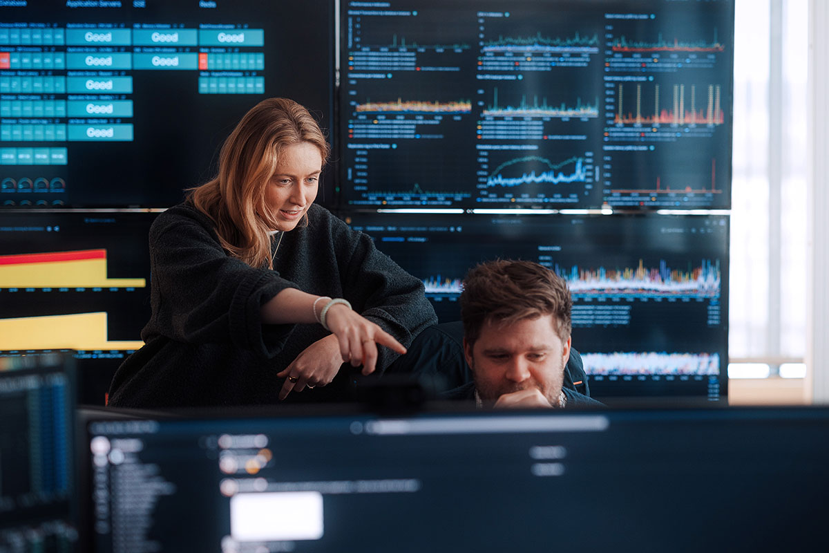 To personer i et kontrollrom overvåker flere dataskjermer. En person peker på en monitor, diskuterer prosjektdetaljer og hjelper den andre bli kjent med Tripletex, mens den andre fokuserer intenst på skjermen sin.