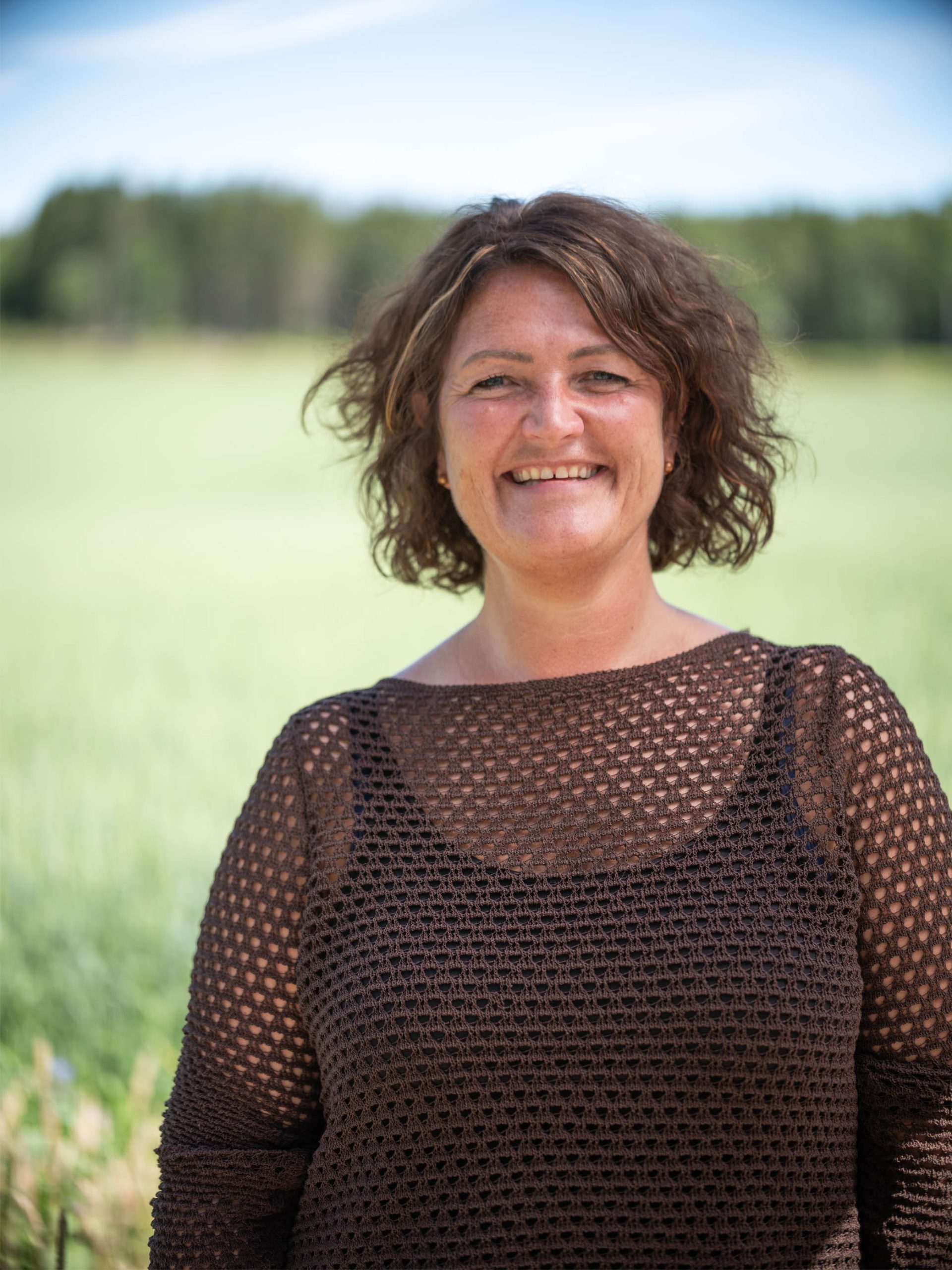 En kvinne med krøllete hår smiler mens hun står på et felt. Hun har på seg en mørkebrun heklet topp.