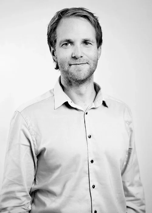 Et svart-hvitt-bilde av en mann med middels langt hår og skjegg, iført en lys button-down skjorte, snakker om Sticos Oversikt.