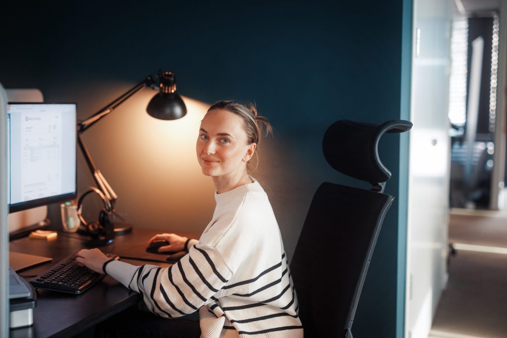 dame med oppsatt hår og stripete genser smiler til kamera mens hun sitter og jobber på pcen