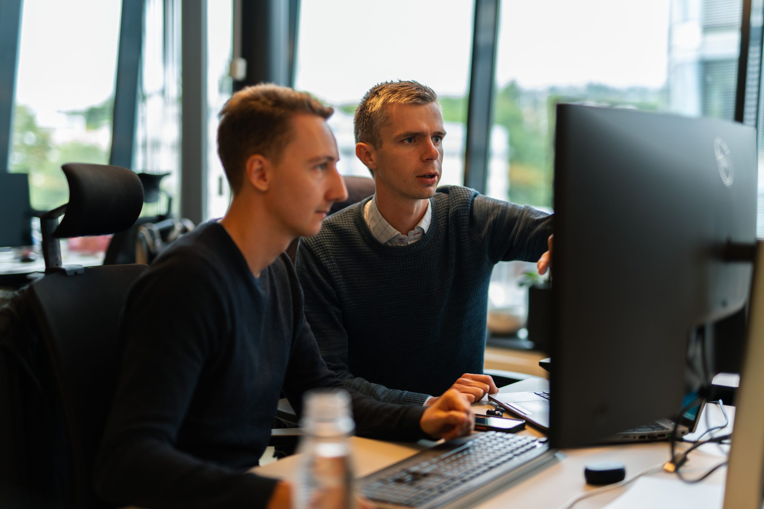 To menn sitter ved et skrivebord foran en dataskjerm; den ene peker på skjermen mens den andre følger oppmerksomt med.