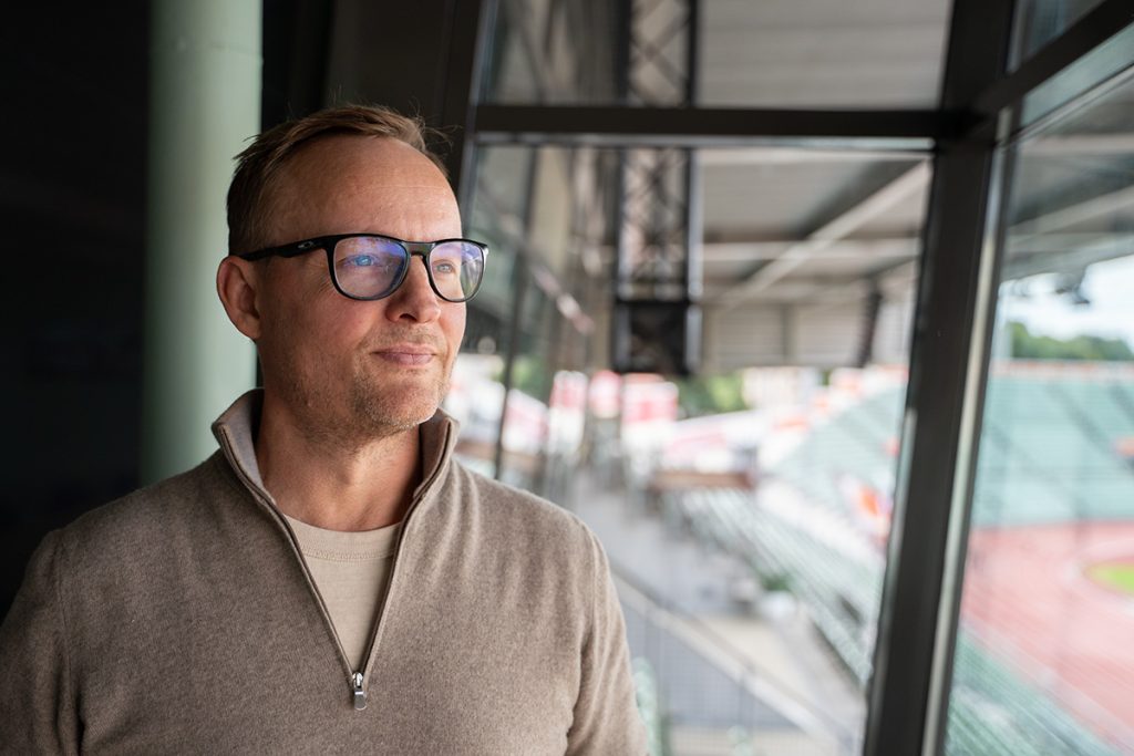 Stian Andersen har på briller og beige genser ser ut gjennom et stort vindu over stadion, der Tinde har lokaler.