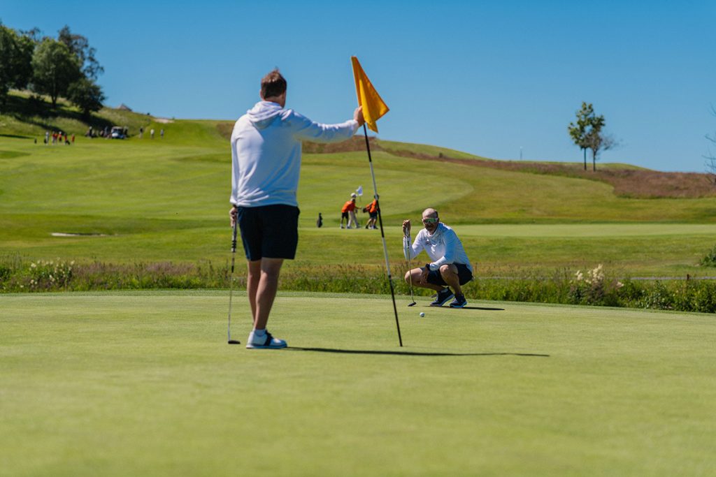 To golfere er på en puttinggreen; den ene holder flaggstangen mens den andre stiller opp en putt.