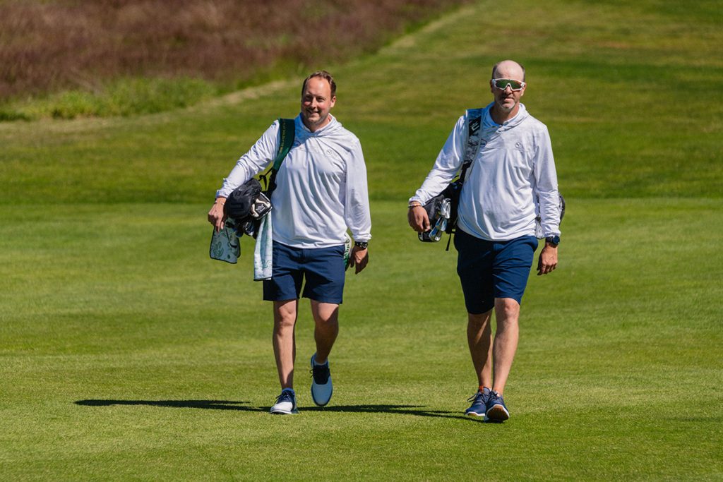 To menn i matchende hvite skjorter og marine shorts går over en gresskledd golfbane, med hver sitt sett med golfkøller.