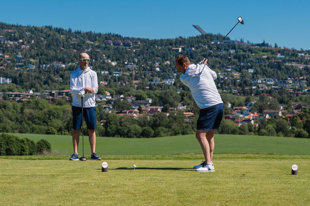 To menn er på en golfbane, den ene svinger en golfkølle mens den andre ser på.