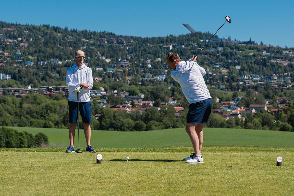 To menn er på en golfbane, den ene svinger en golfkølle mens den andre ser på.