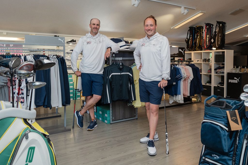 To menn i matchende atletiske antrekk står i en butikk fylt med golfutstyr og klær. Den ene lener seg på et klesstativ, og den andre holder en golfkølle.