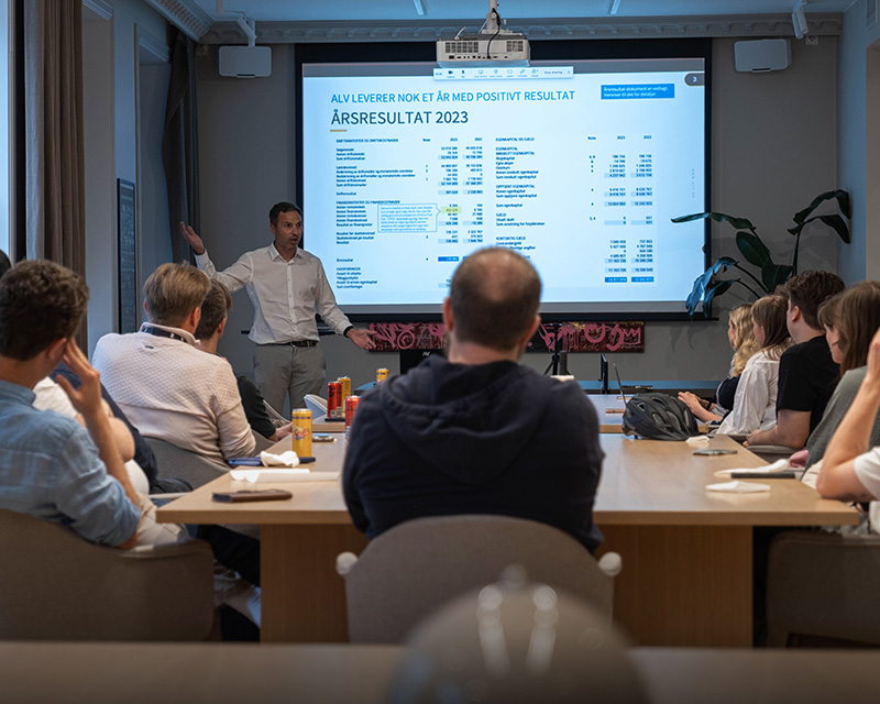 En person presenterer økonomiske resultater på en skjerm for en sittende gruppe i et konferanserom.