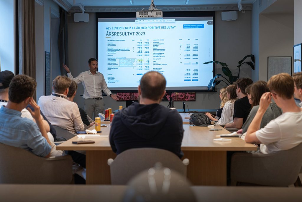 En person presenterer økonomiske resultater på en skjerm for en gruppe som sitter rundt et konferansebord.