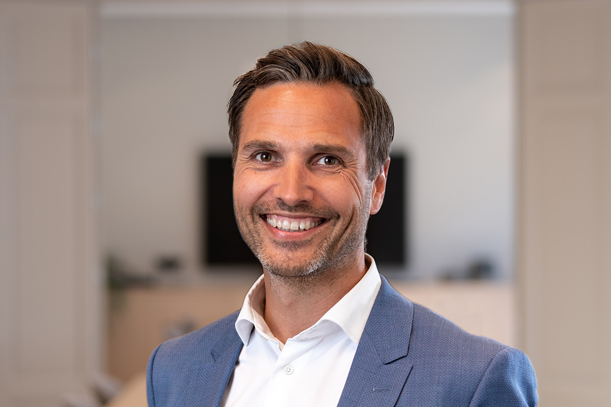En smilende mann i blå blazer og hvit skjorte står i kontormiljø. Marcus fra Alv AS