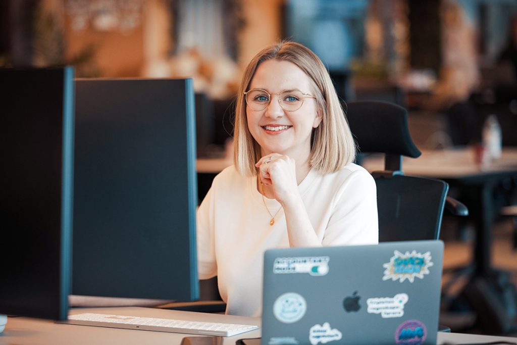Hanne Lundby smiler ved et skrivebord, glad for at vi er medlem av oda-nettverk, med en dataskjerm og en klistremerkebelagt bærbar PC.