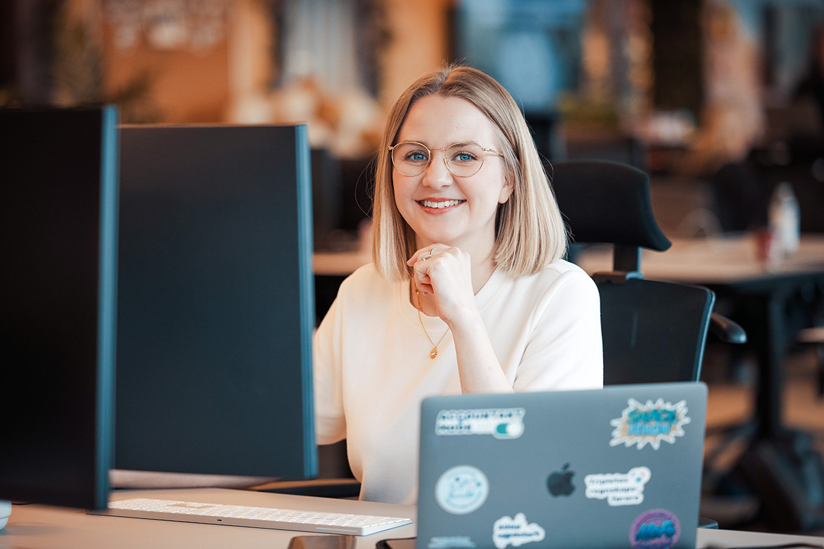 Hanne Lundby smiler ved et skrivebord, glad for at vi er medlem av oda-nettverk, med en dataskjerm og en klistremerkebelagt bærbar PC.