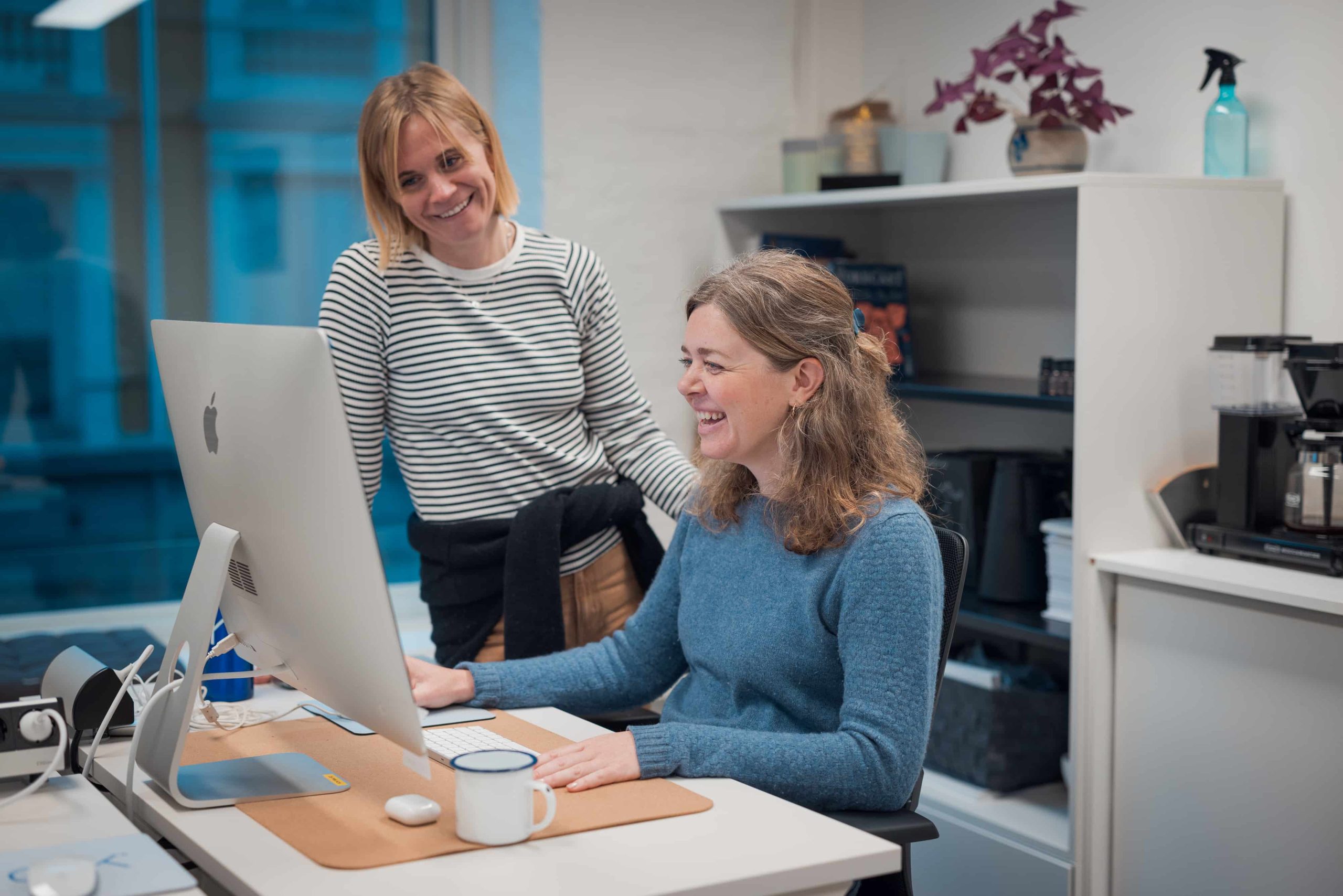 To kvinner på kontor, den ene sitter og jobber ved en datamaskin mens den andre står i nærheten, begge smiler.