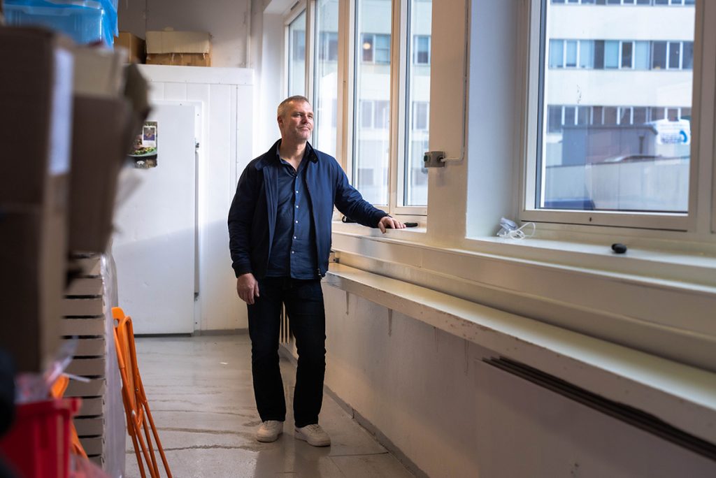 En person står i et rom ved siden av store vinduer og ser utover. De har på seg en mørk jakke, jeans og hvite sko. Rommet har noen bokser og en oransje stol.