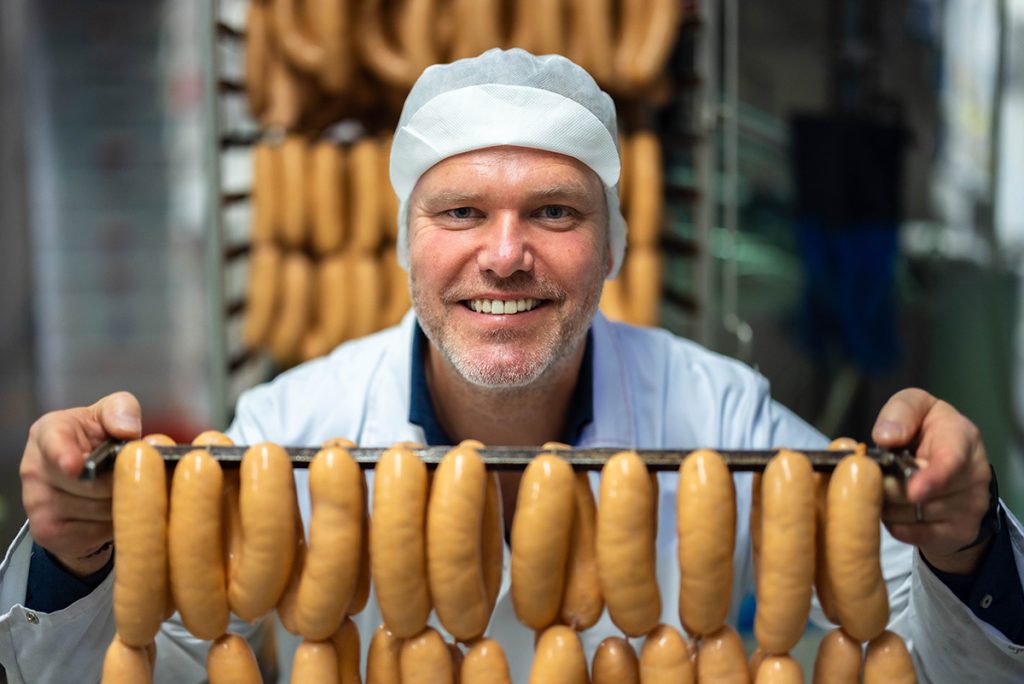 Jon Holm, daglig leder i Metervare, smiler mens han holder et stativ med pølser, iført en hvit frakk og hårnett.
