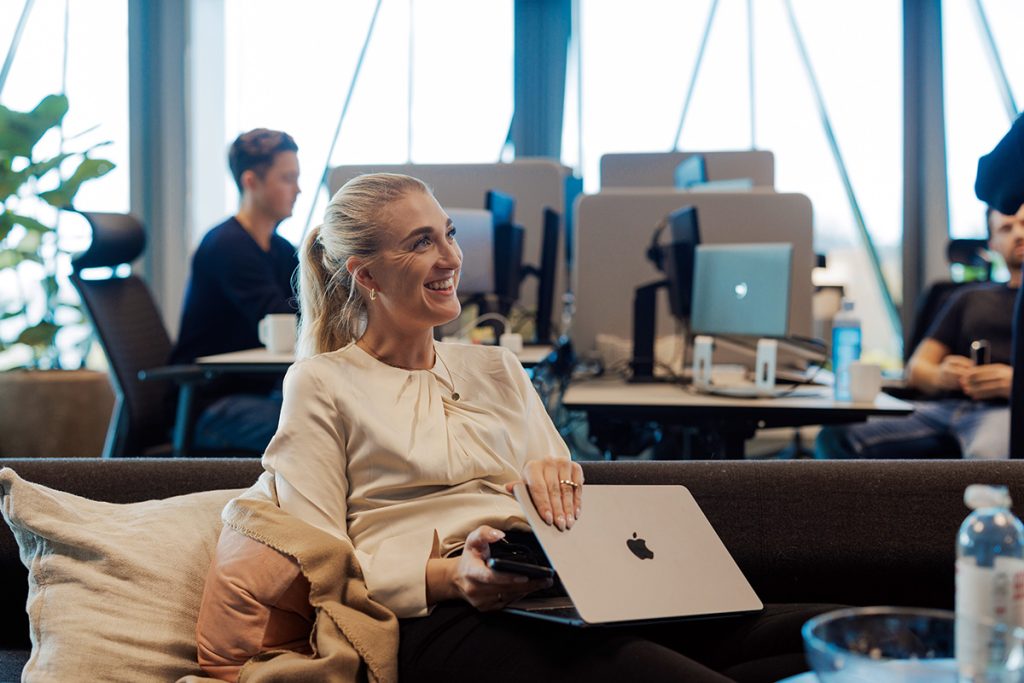 Kvinne sitter på en sofa med en bærbar datamaskin og jobber med forretningsplan. Kontormiljø med folk som jobber bakgrunnen.