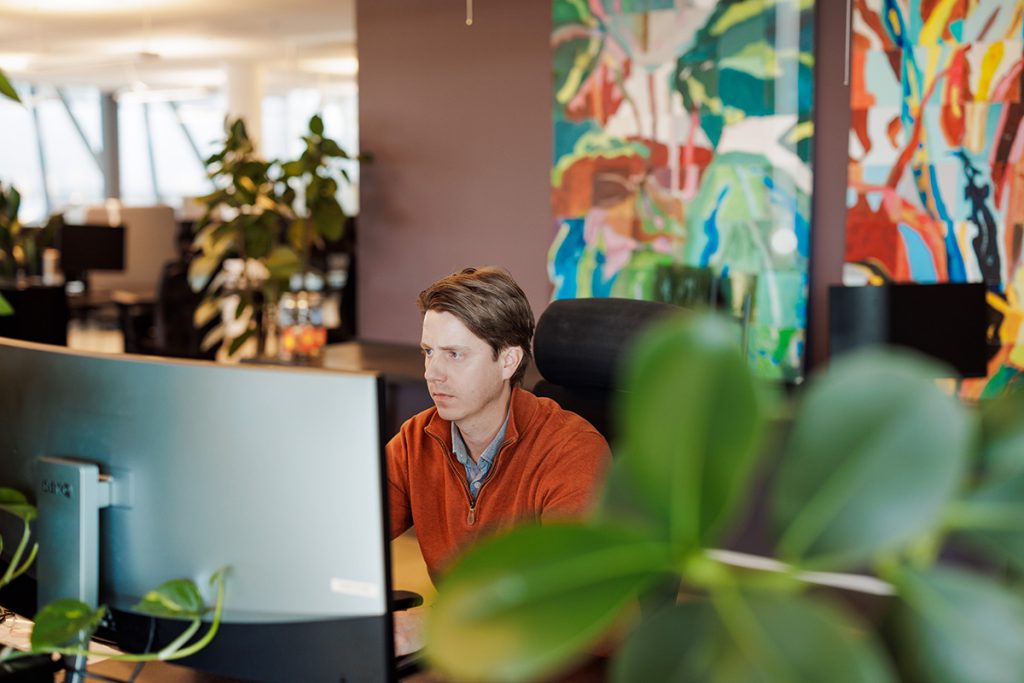En person iført en oransje genser jobber ved en datamaskin på et moderne kontor med abstrakt kunst og planter i bakgrunnen.
