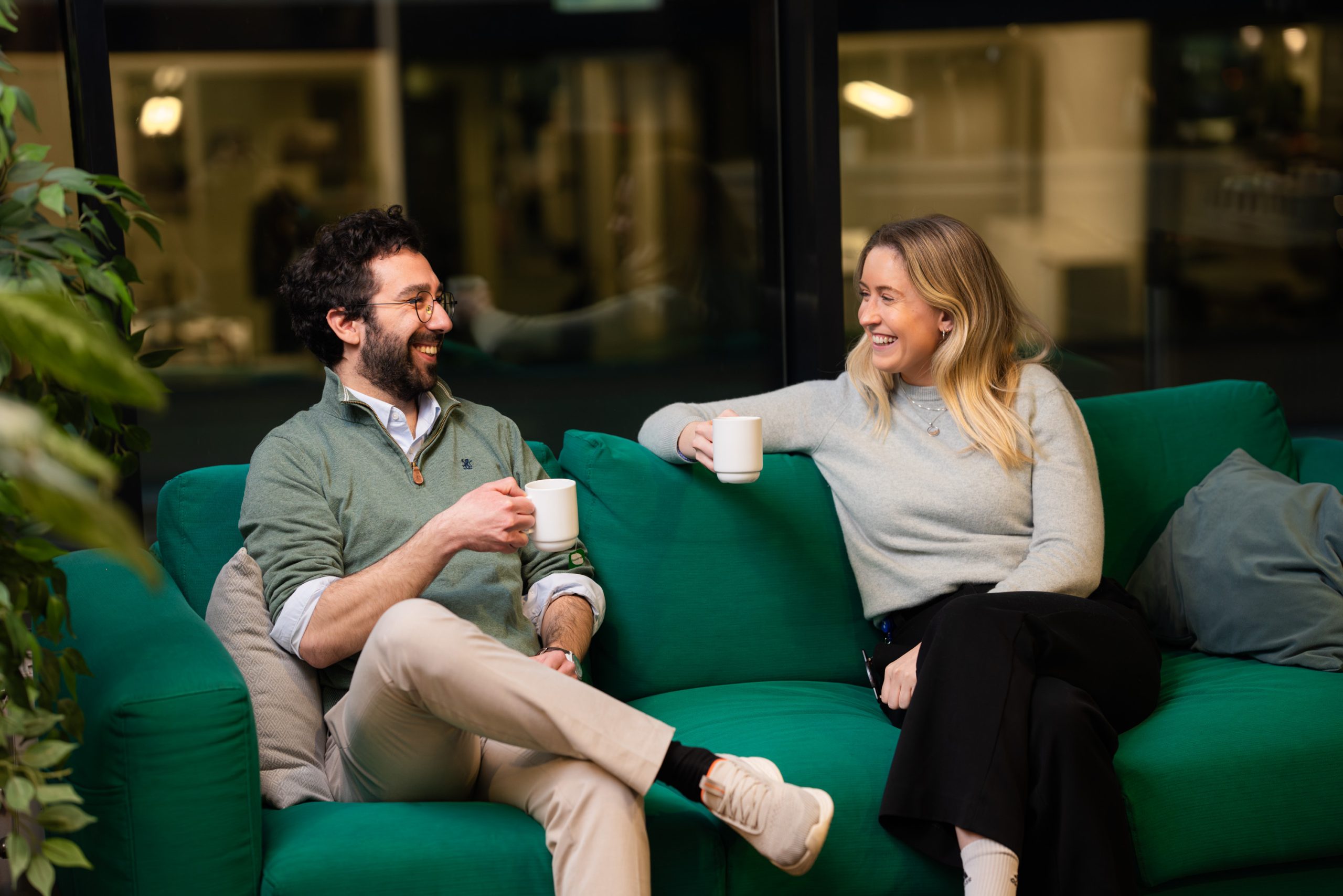 To personer sitter i en grønn sofa, smilende og med hvite krus i hendene, i samtale i et moderne innemiljø.