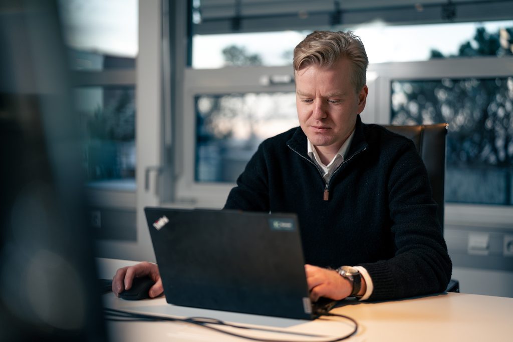 En mann sitter ved et skrivebord og bruker en bærbar datamaskin på et kontor med store vinduer.