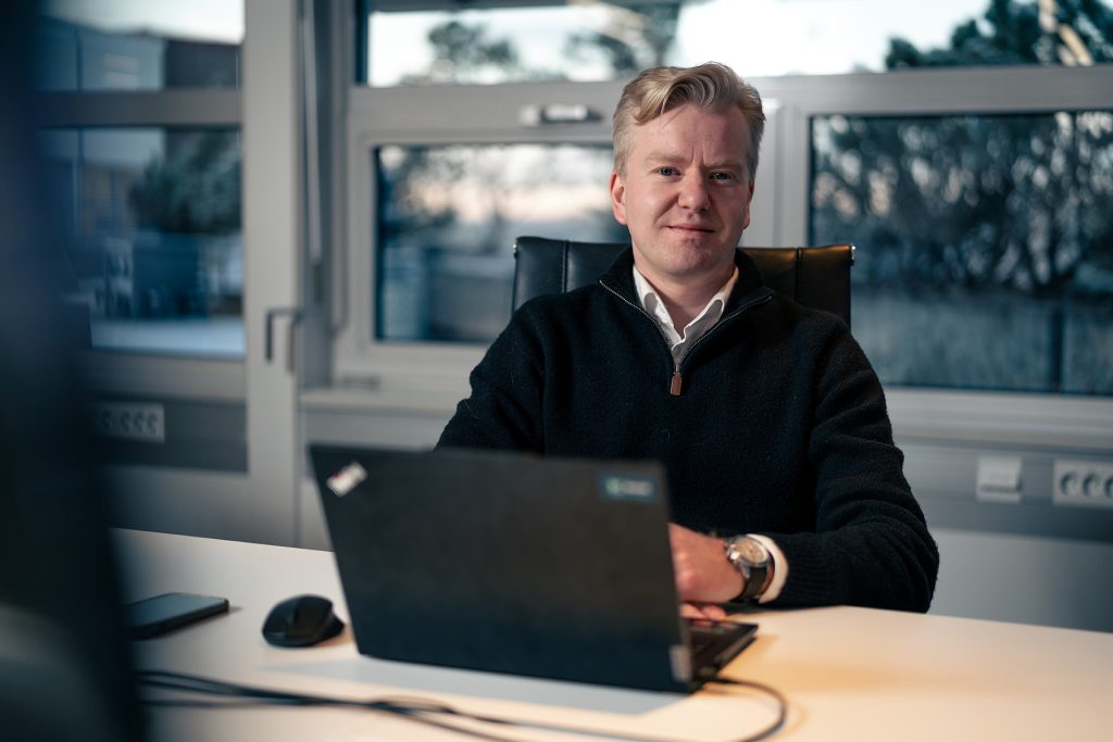 Tobias Aardal i Nordic Infrastructure Group sitter ved et skrivebord med en åpen bærbar PC, iført en mørk genser. Snakker om bærekraft.