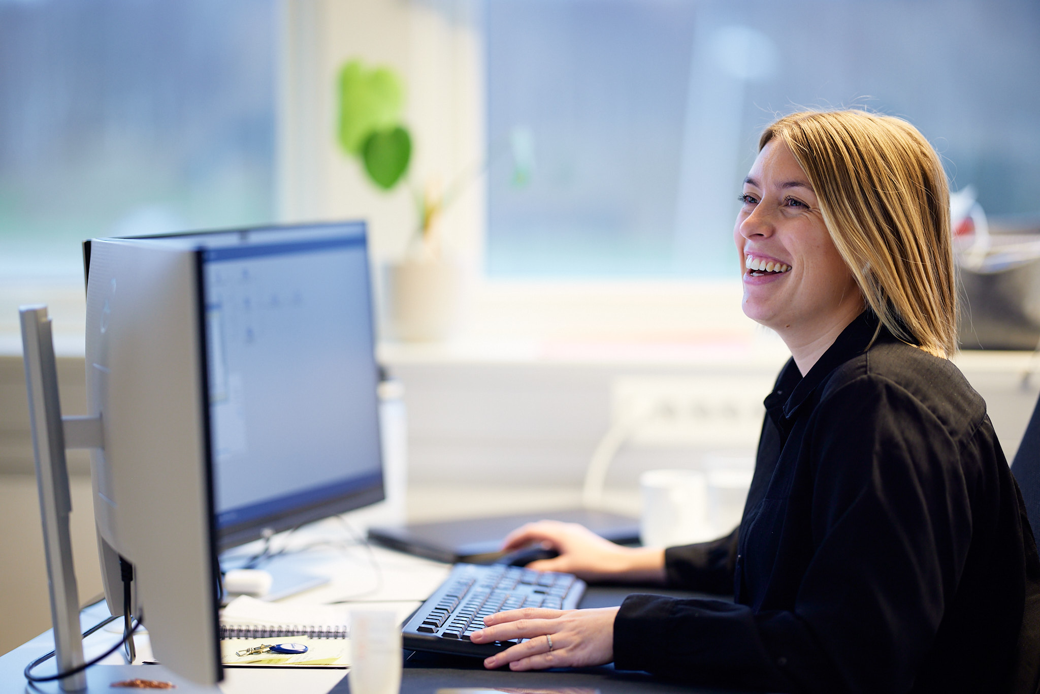En person som sitter ved et skrivebord, smiler og ser på en dataskjerm med Sticos Oversikt.