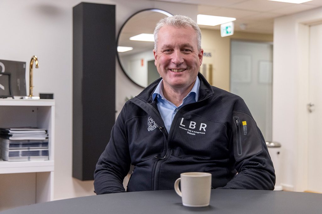 Smilende mann i svart LBR-jakke sitter ved et bord med et krus, i moderne kontormiljø.