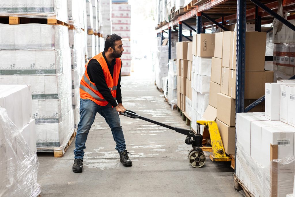 En arbeider i en refleksvest bruker en pallejekk til å flytte esker i en lagergang, og administrerer varelageret effektivt med Rackbeat og Tripletex.