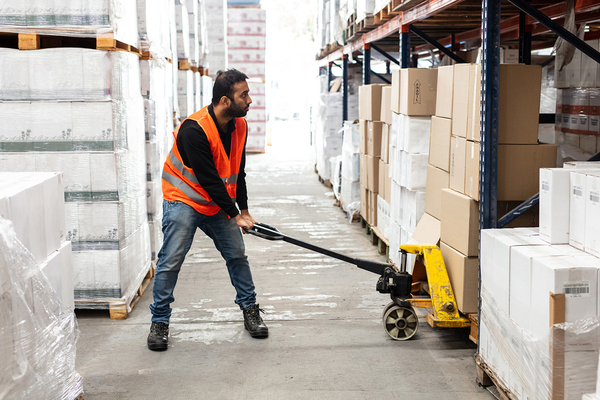 En arbeider i en refleksvest bruker en pallejekk til å flytte esker i en lagergang, og administrerer varelageret effektivt med Rackbeat og Tripletex.