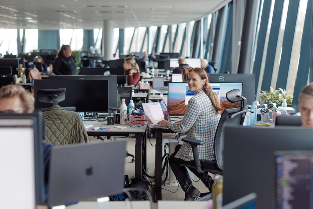 Åpent kontorlandskap med flere personer som jobber ved skrivebord; én person i midten, sittende og ser inn i kameraet, klart til å sende fakturaer med bare noen få klikk.