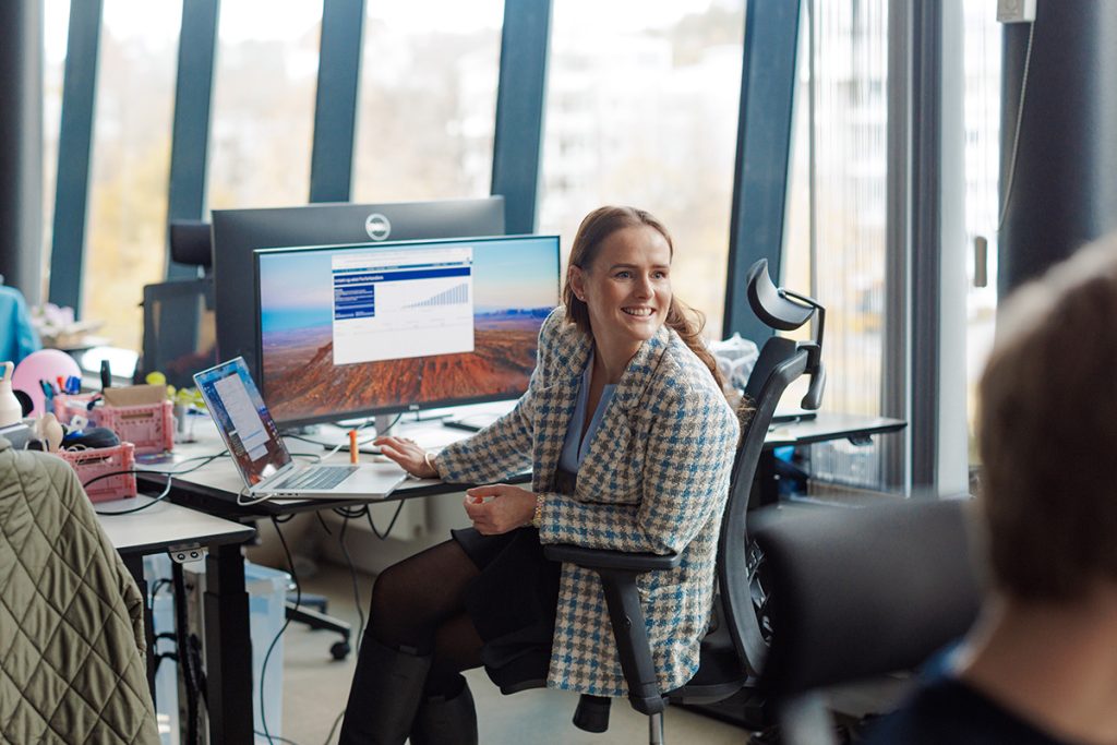 En kvinne sitter ved skrivebordet sitt i et moderne kontor, smiler og snakker med en kollega, med en bærbar PC og en stor skjerm som viser en graf mens hun forbereder seg på å sende faktura.