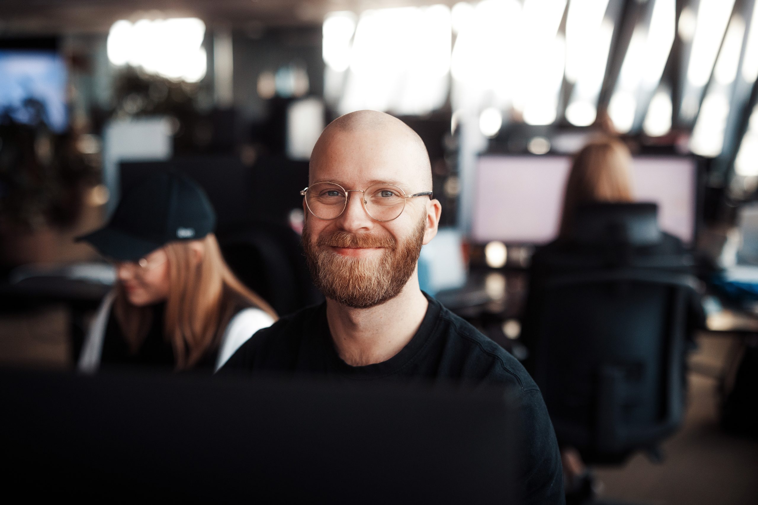 En skjeggete mann med briller smiler til kameraet i moderne kontormiljø, mens en person med caps jobber i bakgrunnen.