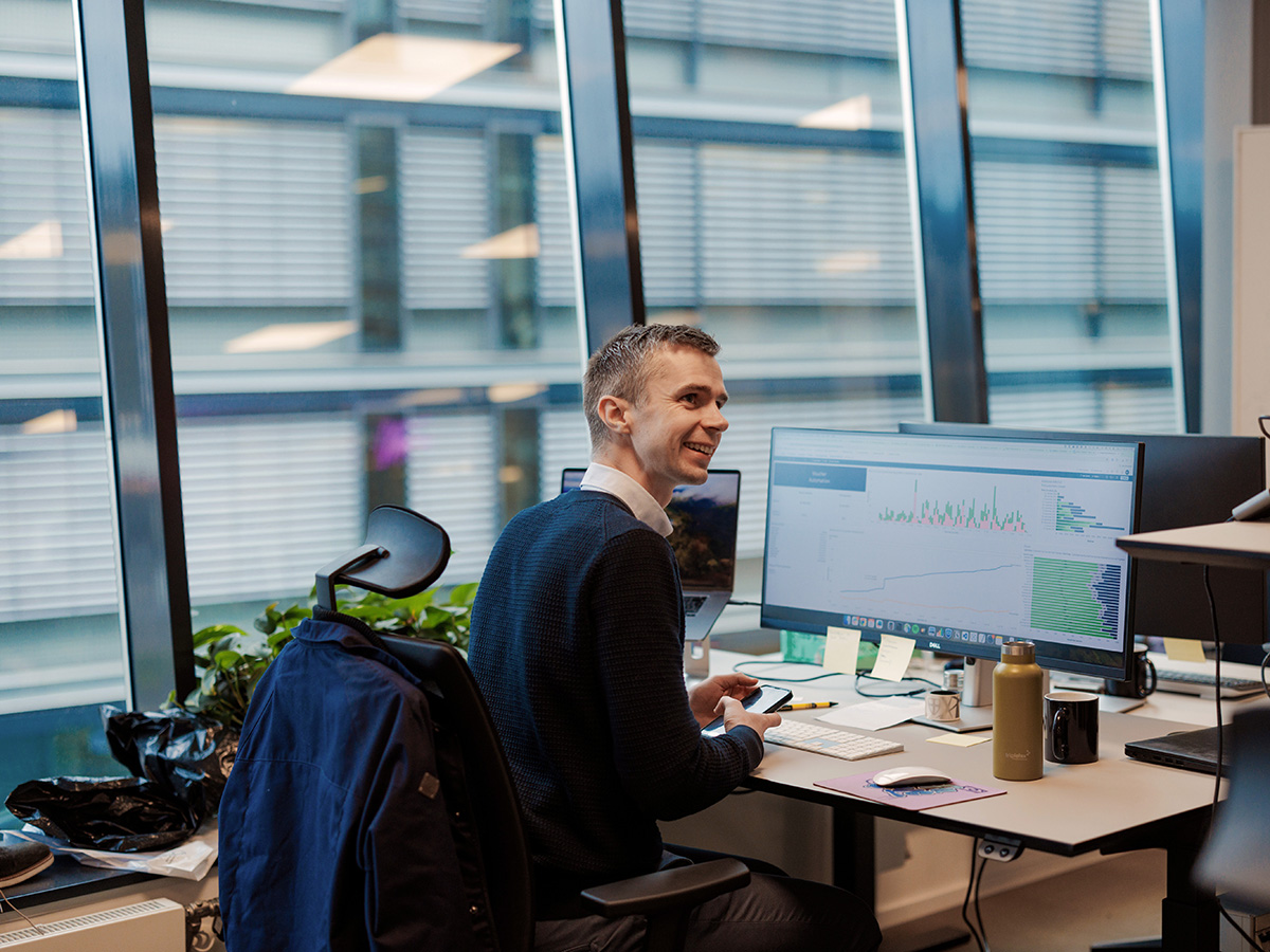 En mann i blå genser sitter ved et skrivebord på et kontor og analyserer aksjonærregisteroppgaven på datamaskinen sin, som viser ulike grafer. Skrivebordet er utsmykket med planter og notater, mens store vinduer gir et lyst bakteppe.
