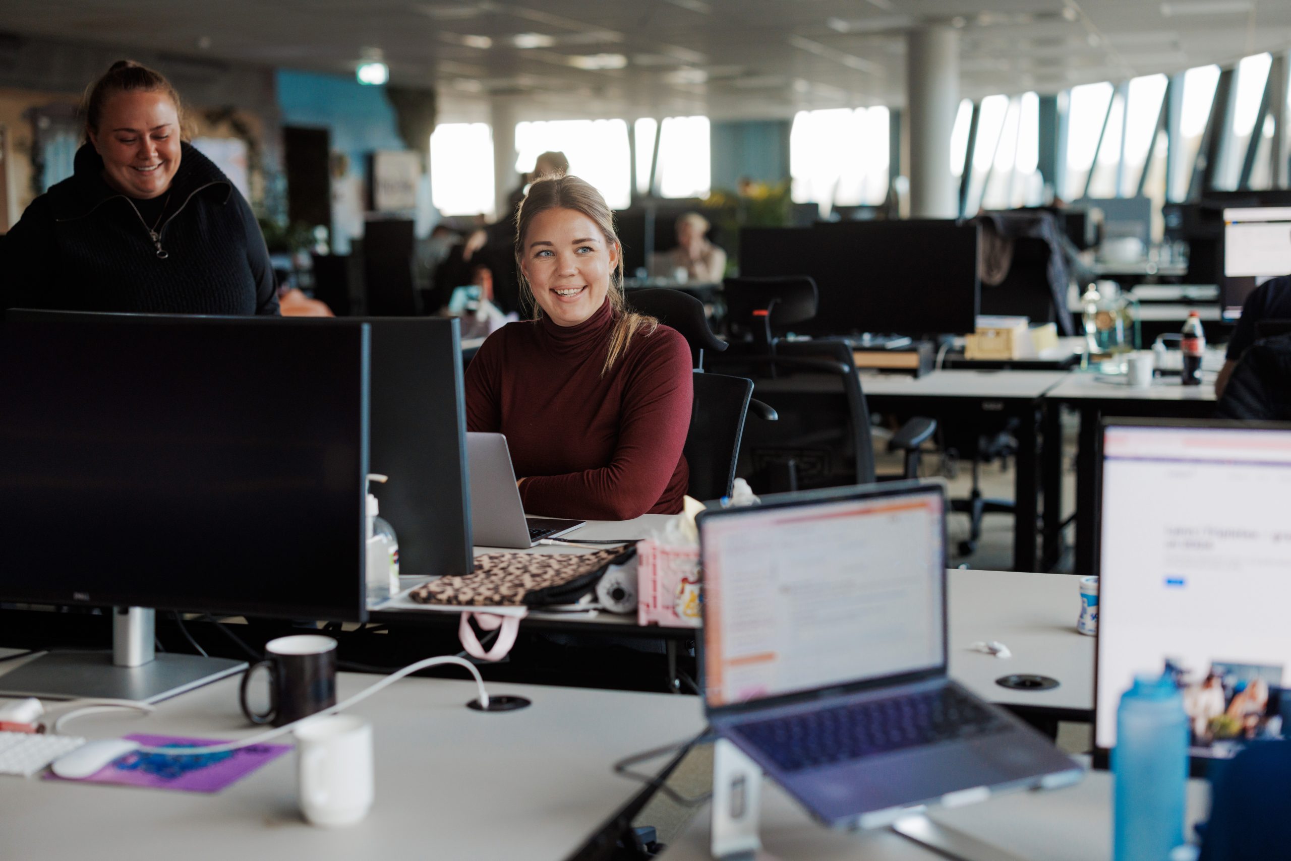 To kvinner samhandler ved et moderne åpent kontorlandskap med datamaskiner, papirer og krus synlige; Anleggsregister Tripletex-programvaren vises på en av skjermene, mens andre pulter og mennesker er i bakgrunnen.