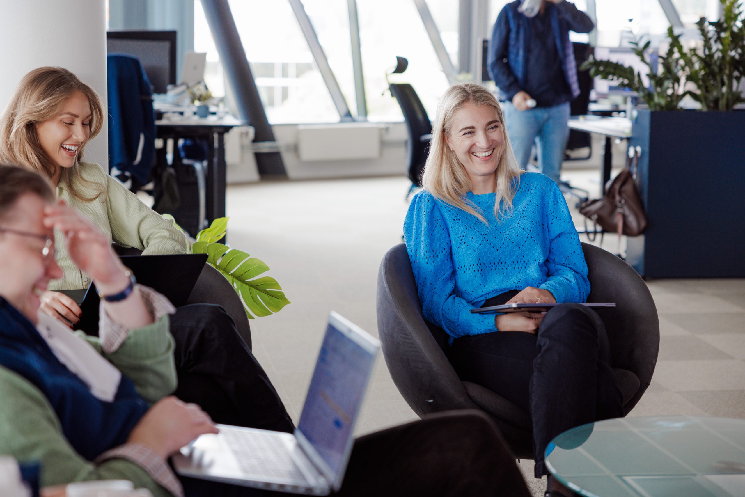 Tre personer sitter i en kontorlounge, to av dem med bærbare datamaskiner, og smiler mens de diskuterer Årsoppgjør kurs; i bakgrunnen skimtes en åpen arbeidsplass og frodige planter.