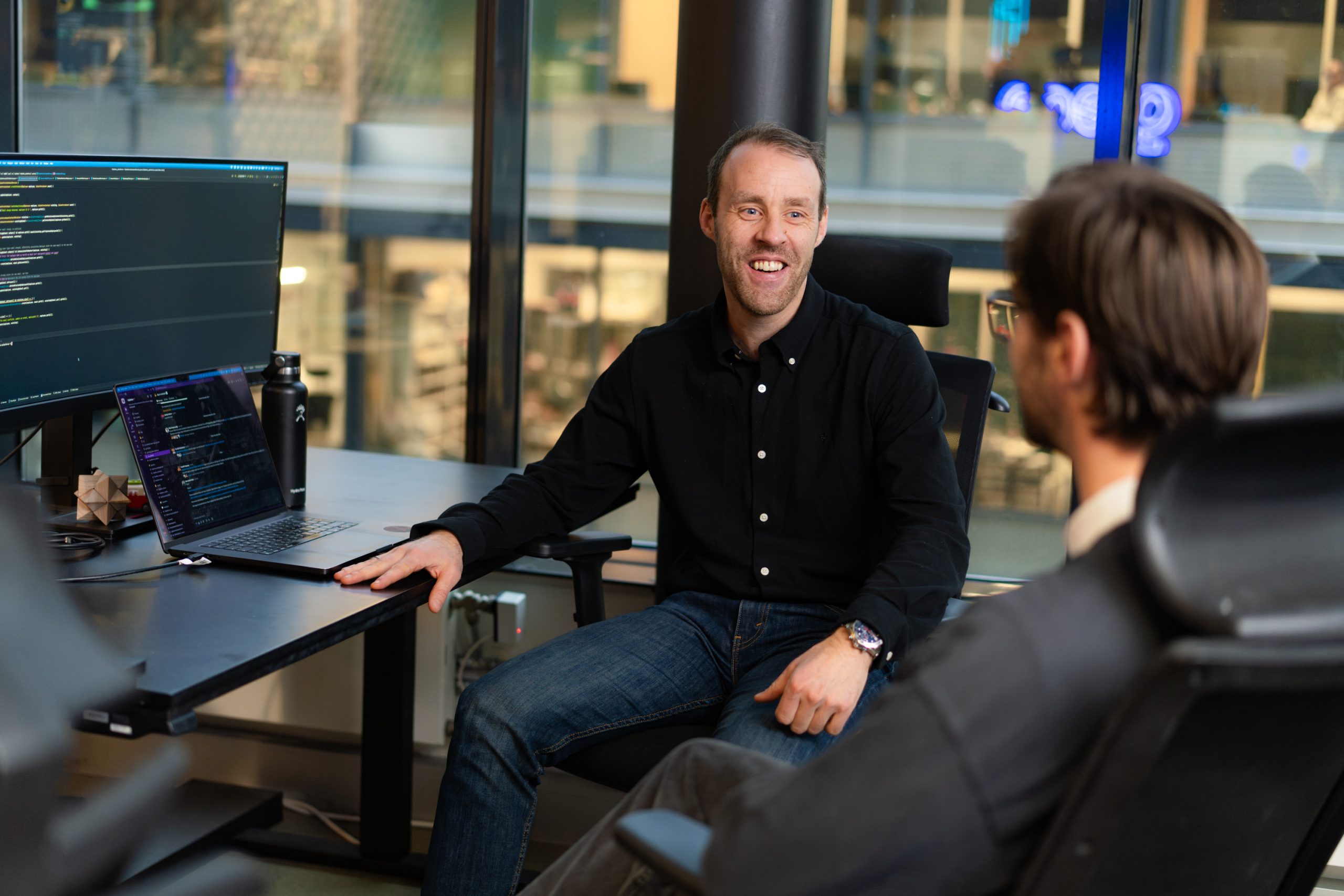 To menn sitter ved skrivebord med dataskjermer som viser kode, og diskuterer et webinar-prosjekt i et moderne kontorlandskap med store vinduer.