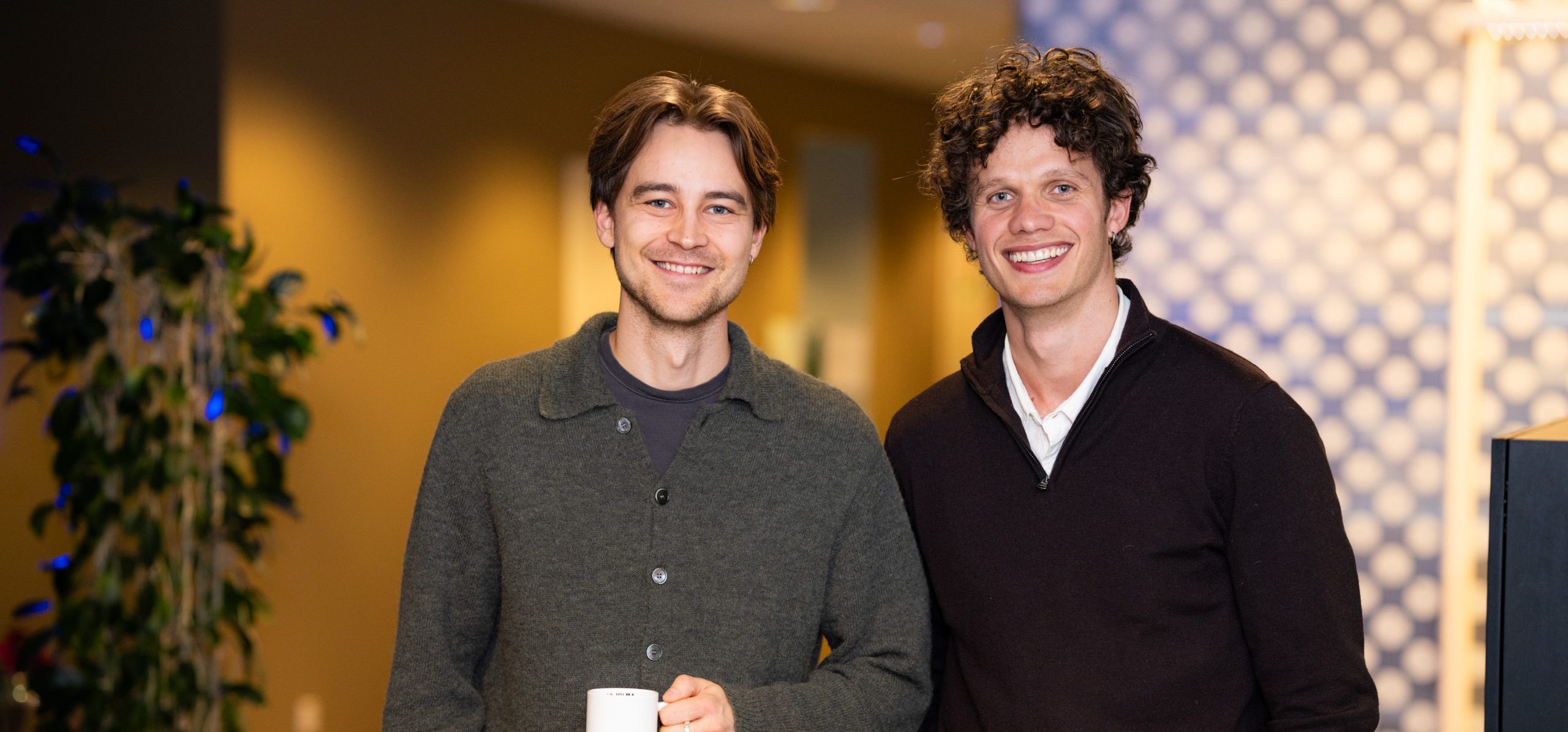 To menn står innendørs og smiler til kameraet; den ene holder et hvitt krus. En potteplante og en mønstret vegg gir sjarm til bakgrunnen. Perfekt for diskusjoner om Årsoppgjør landbruk i en vennlig og profesjonell setting.