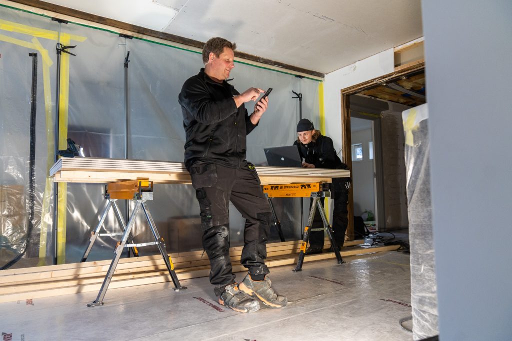 To bygningsarbeidere i et delvis ferdigstilt rom. En fører timer på prosjekt, mens den andre ser på Tripletex på en bærbar datamaskin på en arbeidsbenk.